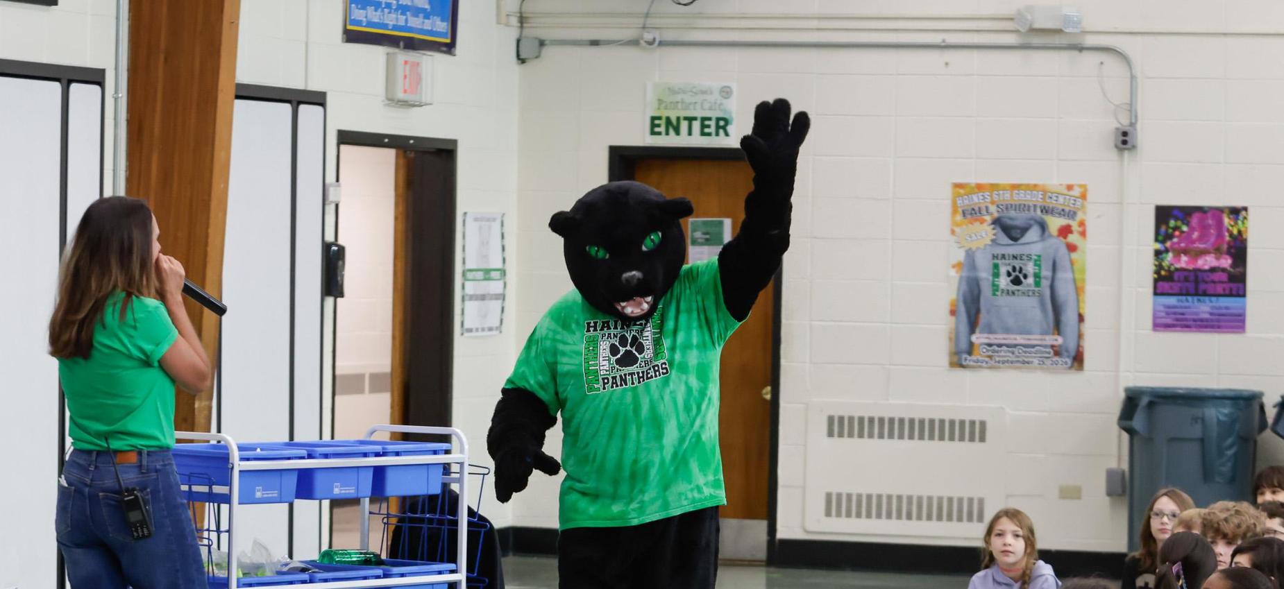 A person in a green shirt speaks to students while a mascot waves.