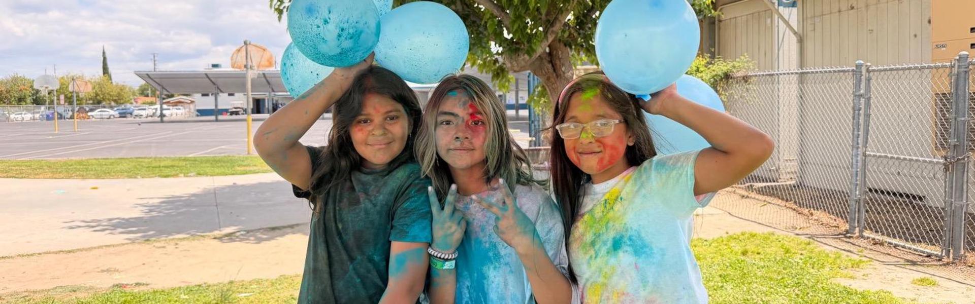 Three girls with colorful faces holding blue balloons, standing under a tree.