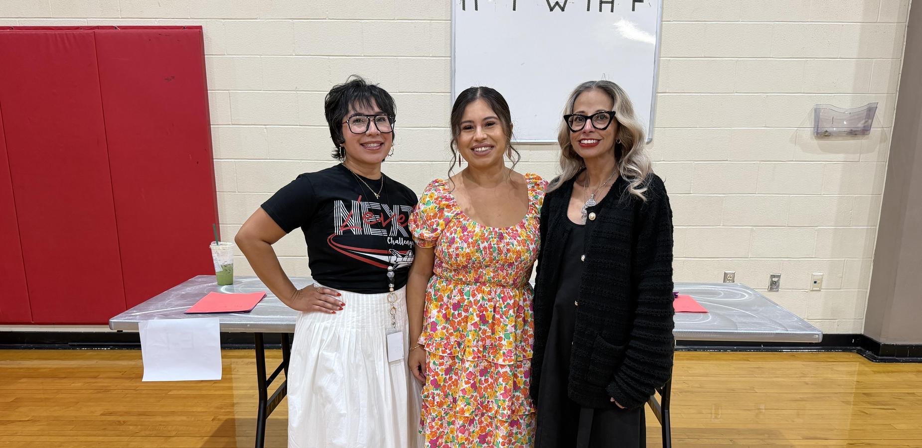 Three women pose together in a gym, wearing stylish outfits and accessories.