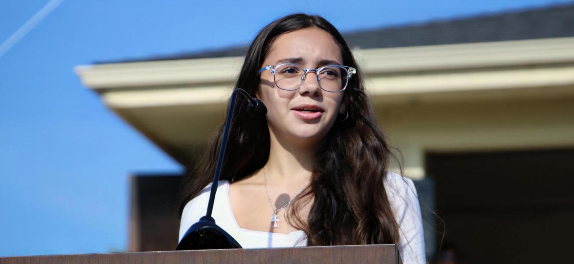 Young woman with glasses speaking at a podium outdoors.
