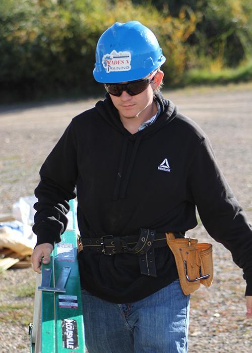 Young man in a blue hard hat carrying a ladder, wearing a black hoodie and tool belt.