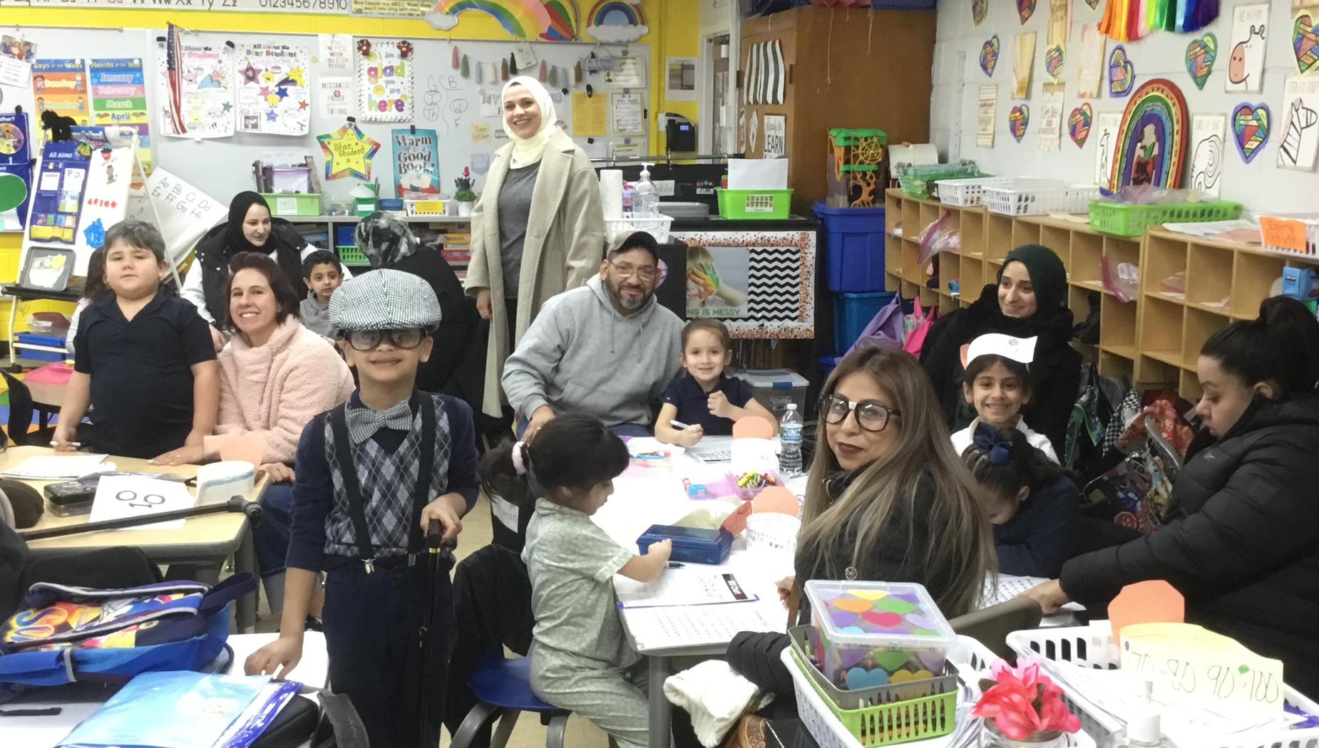 A diverse group of children and adults participate in a classroom activity, some wearing hats.