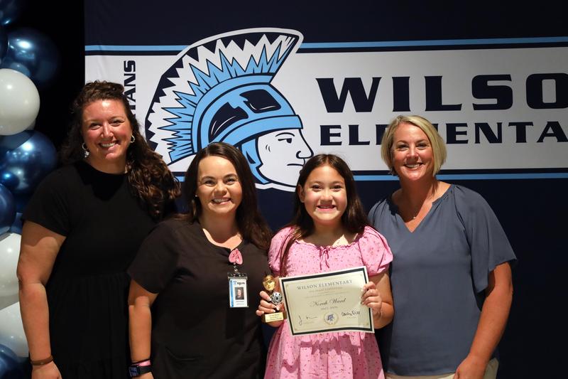 Three women stand with girl in pink dress holding trophy and certificate