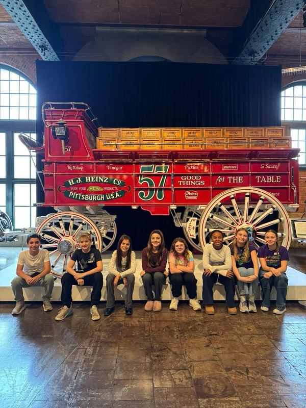 Image depicts a team of Quaker Valley students posing in front of a large, red wagon at the Heinz History Center.
