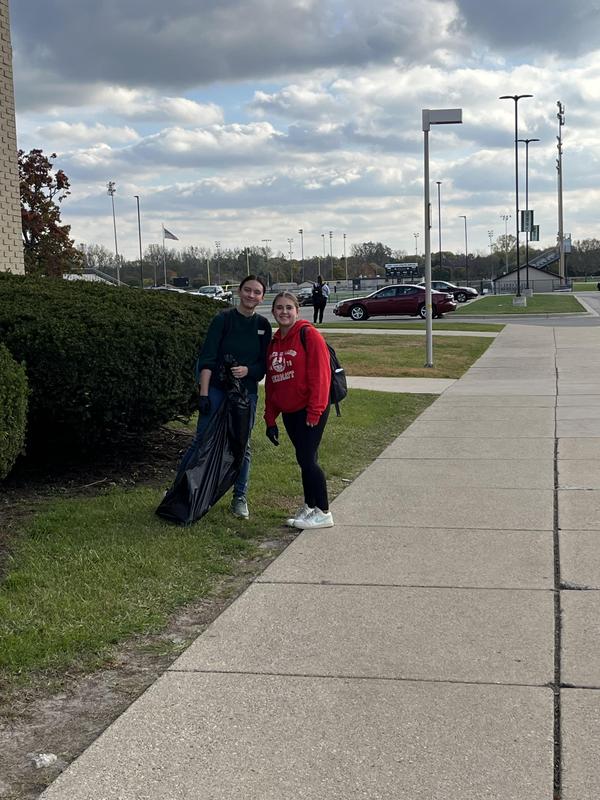 Two girls smiling while holding a garbage bag near a building and grassy area.