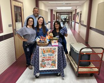 Four adults posing with a cart of snacks and drinks in a school hallway.