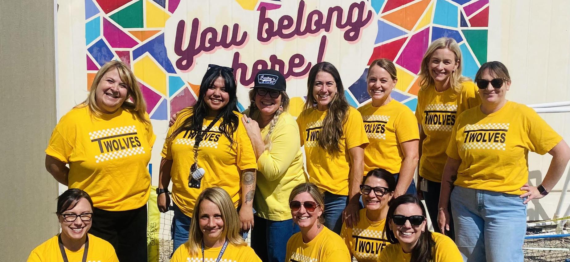 A group of twelve women in matching yellow shirts with 'TWOLVES' printed on them, smiling in front of a colorful mural.