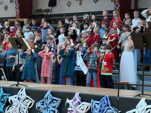 A large group of children perform on stage in festive attire with two microphones in front.
