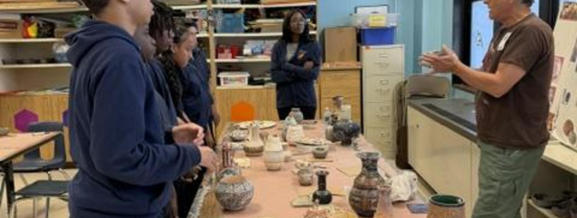 A group of students observing a pottery demonstration in a classroom.