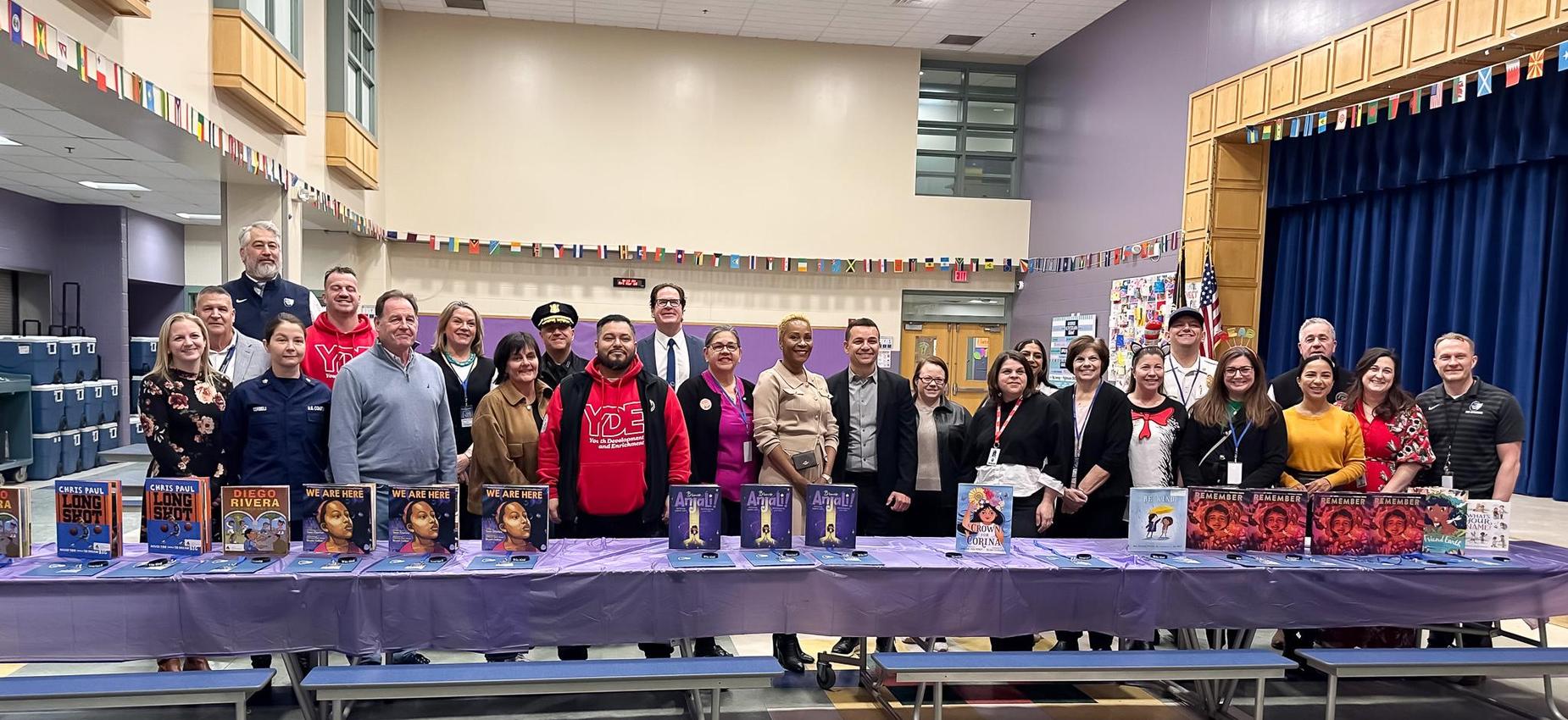 A group of diverse individuals posing together in a school hall, with a display of books on a table.