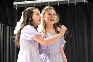 Two girls in lilac dresses singing into a microphone