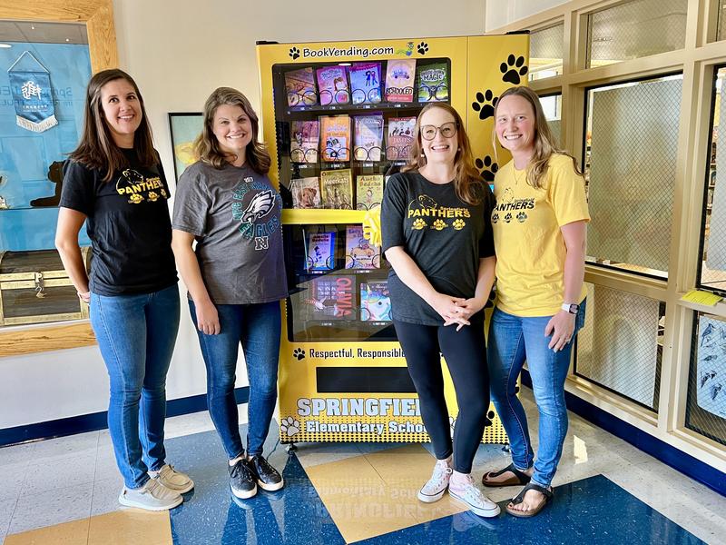 Four PTA officers standing in front of a book vending machine with a ribbon on it.