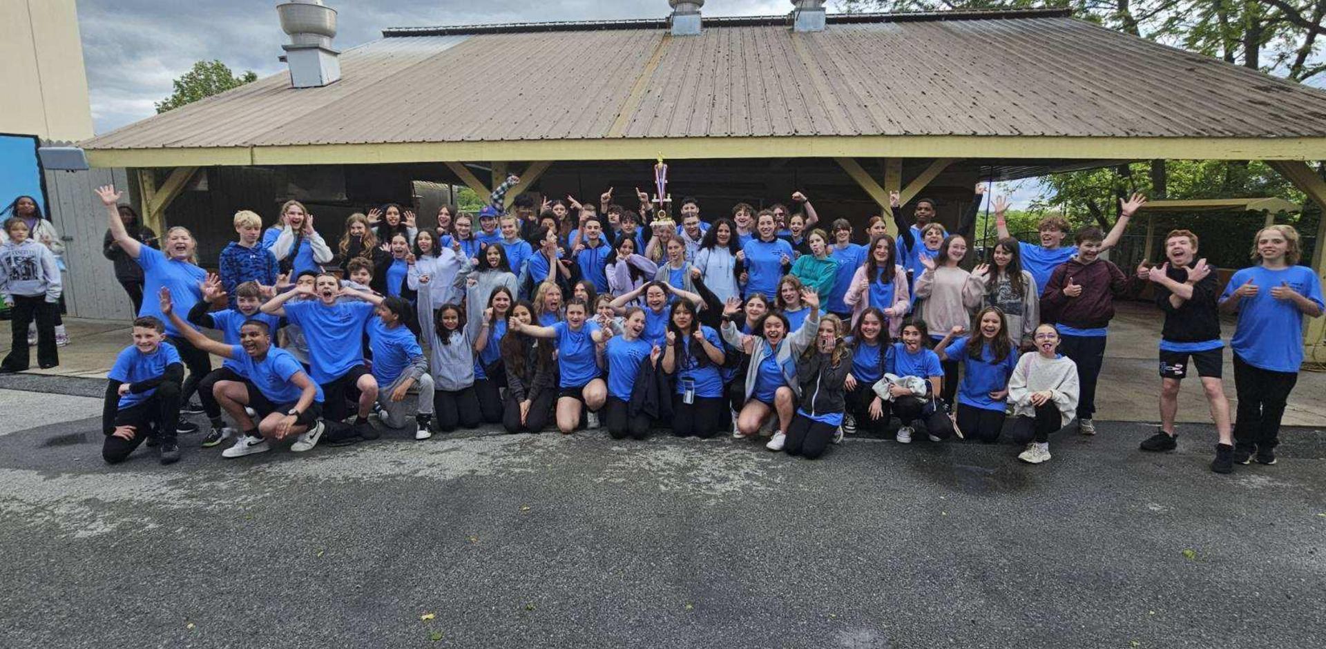 Group of students in blue shirts posing and celebrating outdoors under a shelter.