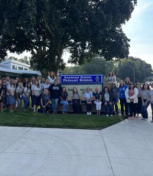 Group of teachers and staff posing in front of a school sign.