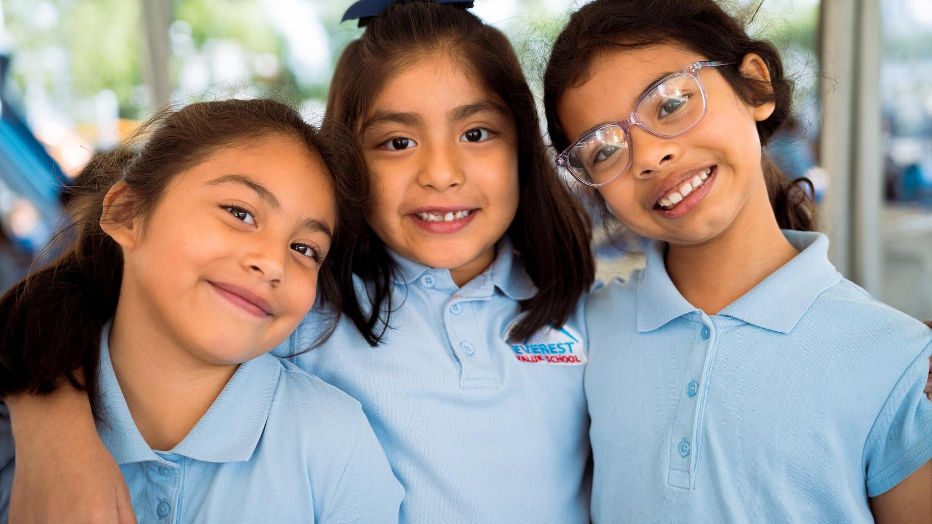 Three girls smiling and posing closely, wearing light blue shirts and glasses.