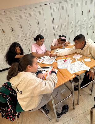 Five students gathered around a table working on arts and crafts with papers and supplies.