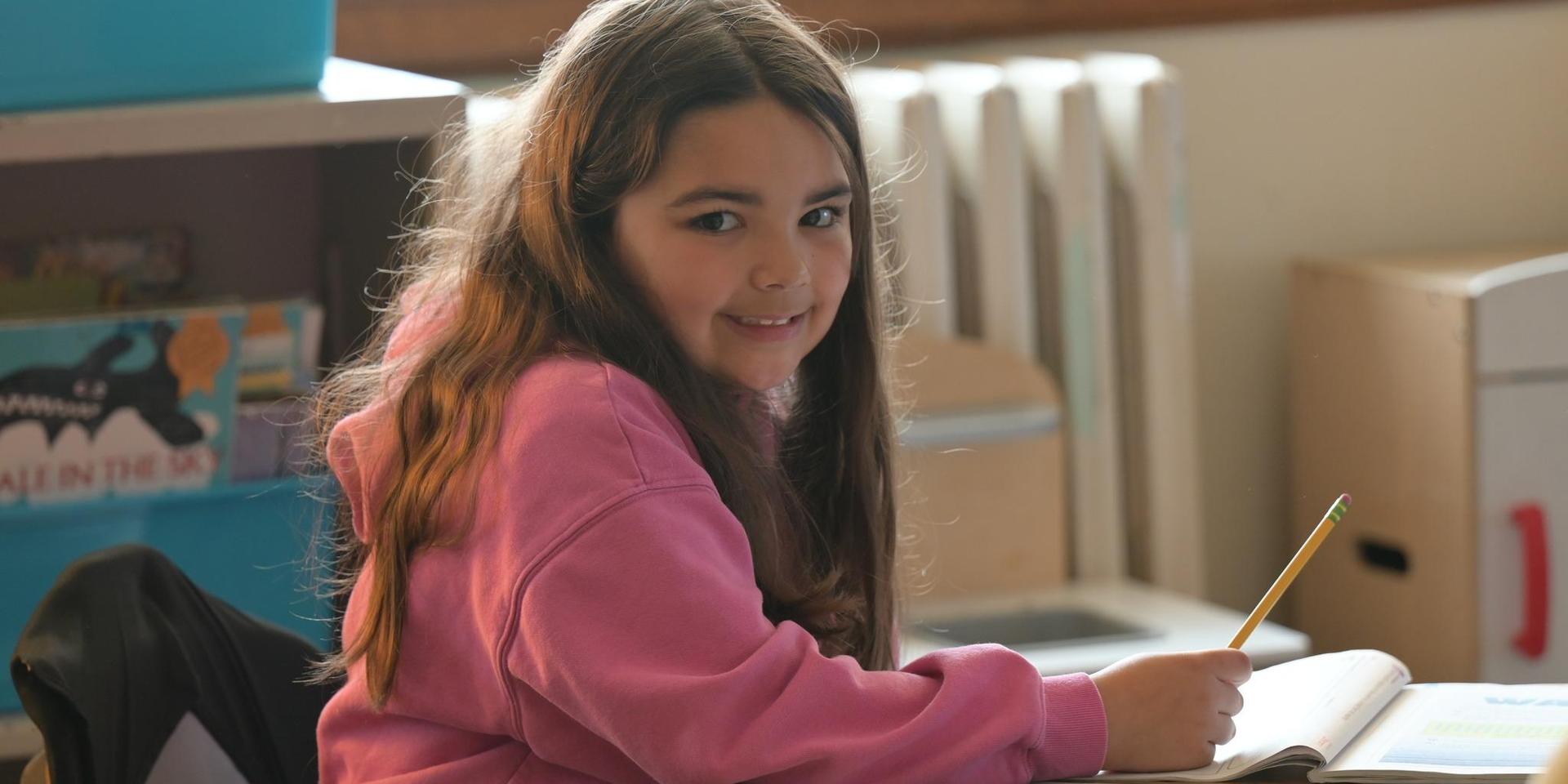 A girl in a pink hoodie smiles while writing in a notebook at her desk.