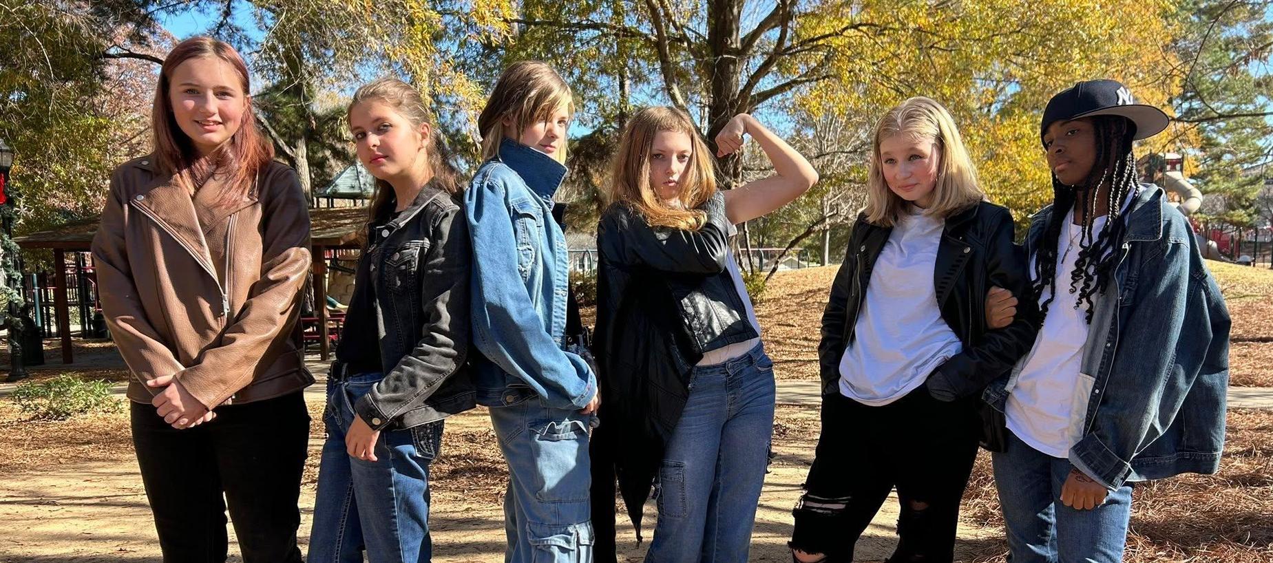 Six young women posing in casual outfits in an outdoor park setting.