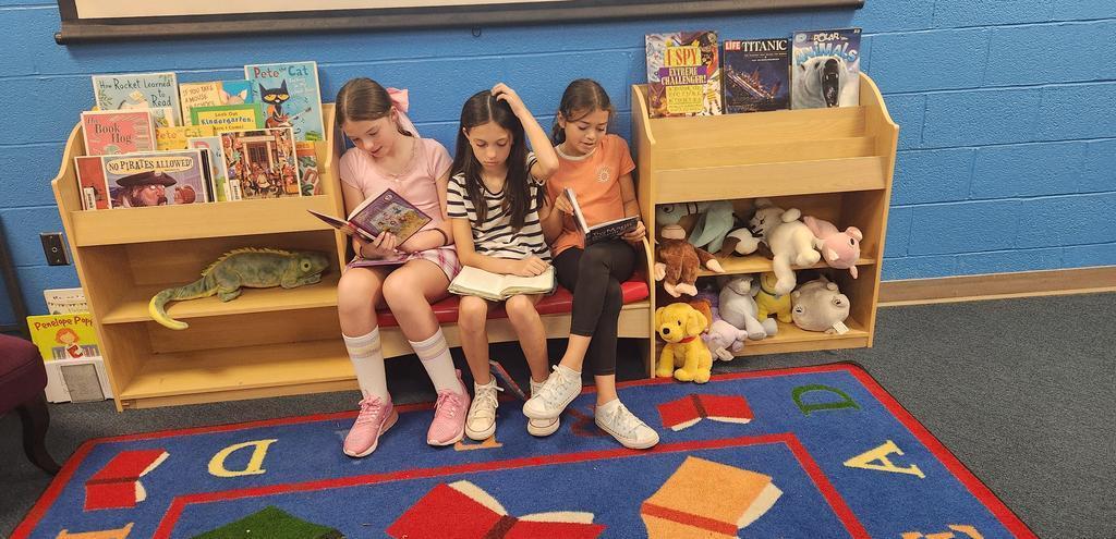 3 students reading on a library bench surrounded by books and stuffed animals