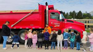 Young students stand next to a large red dump truck while listening to a community volunteer.