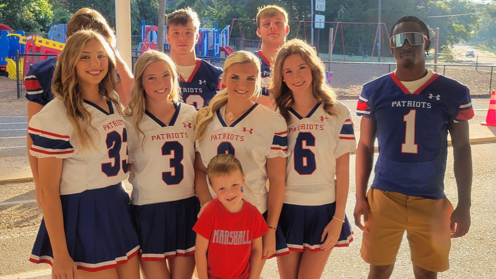 Cheerleaders in uniforms and a boy in red pose together outside a school.