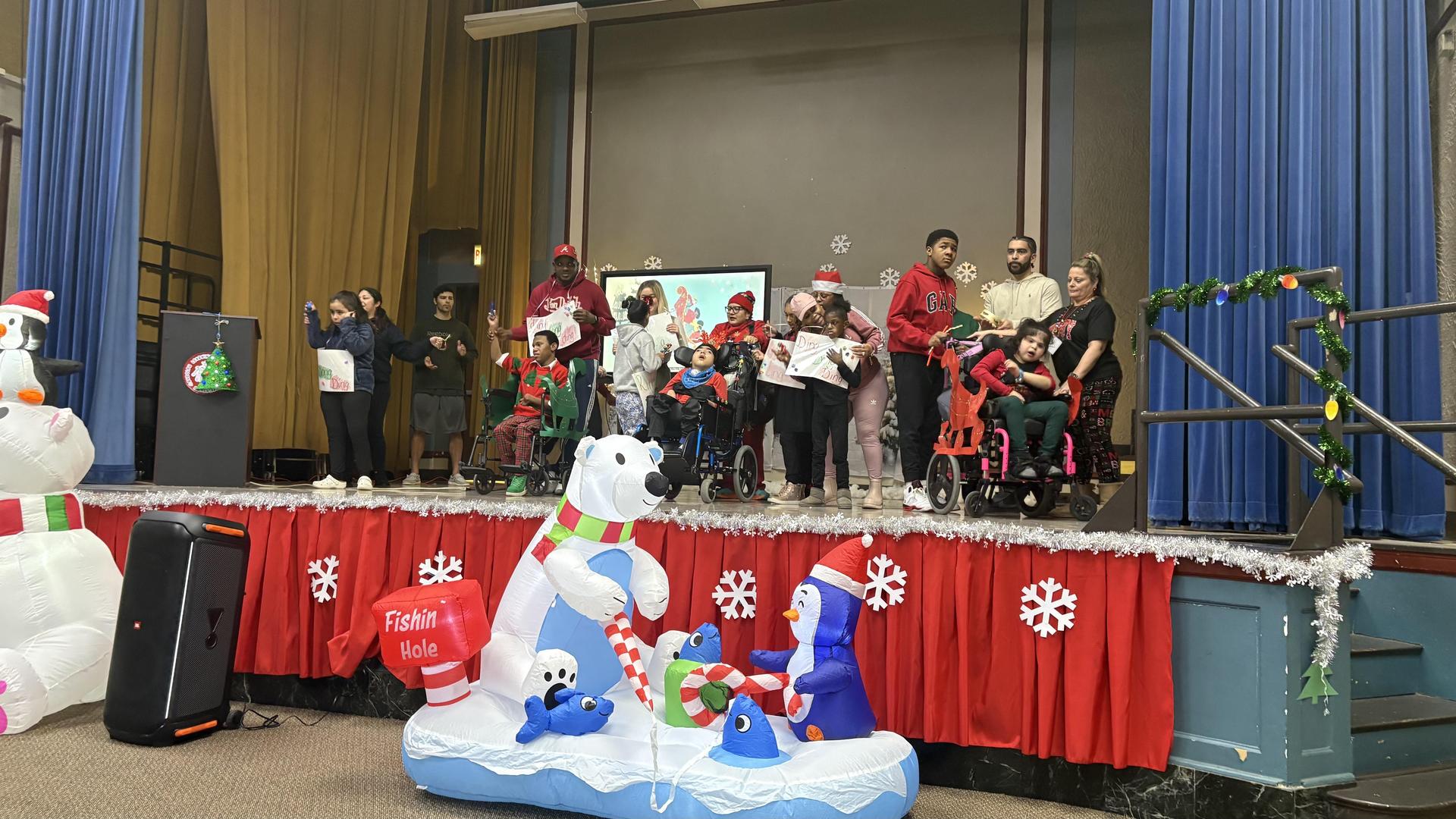 Group of children and adults performing with holiday cards, inflatable polar bear in front.