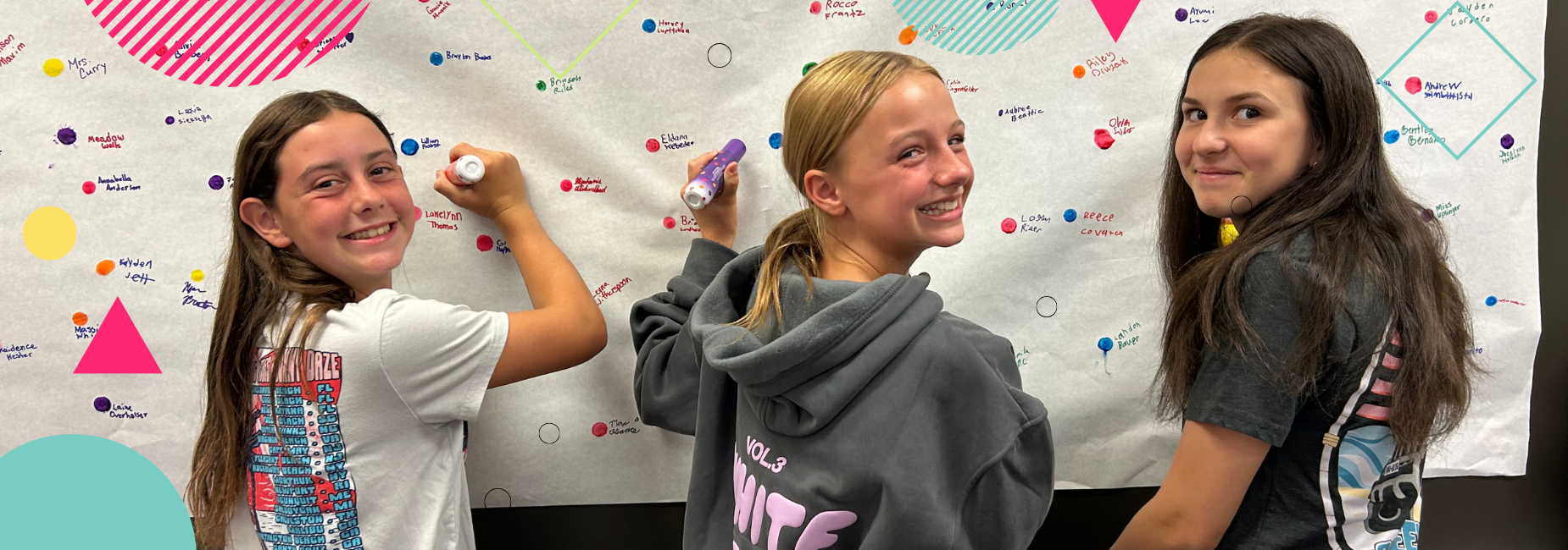 Three girls joyfully writing on a decorated wall covered in colorful dots and designs.