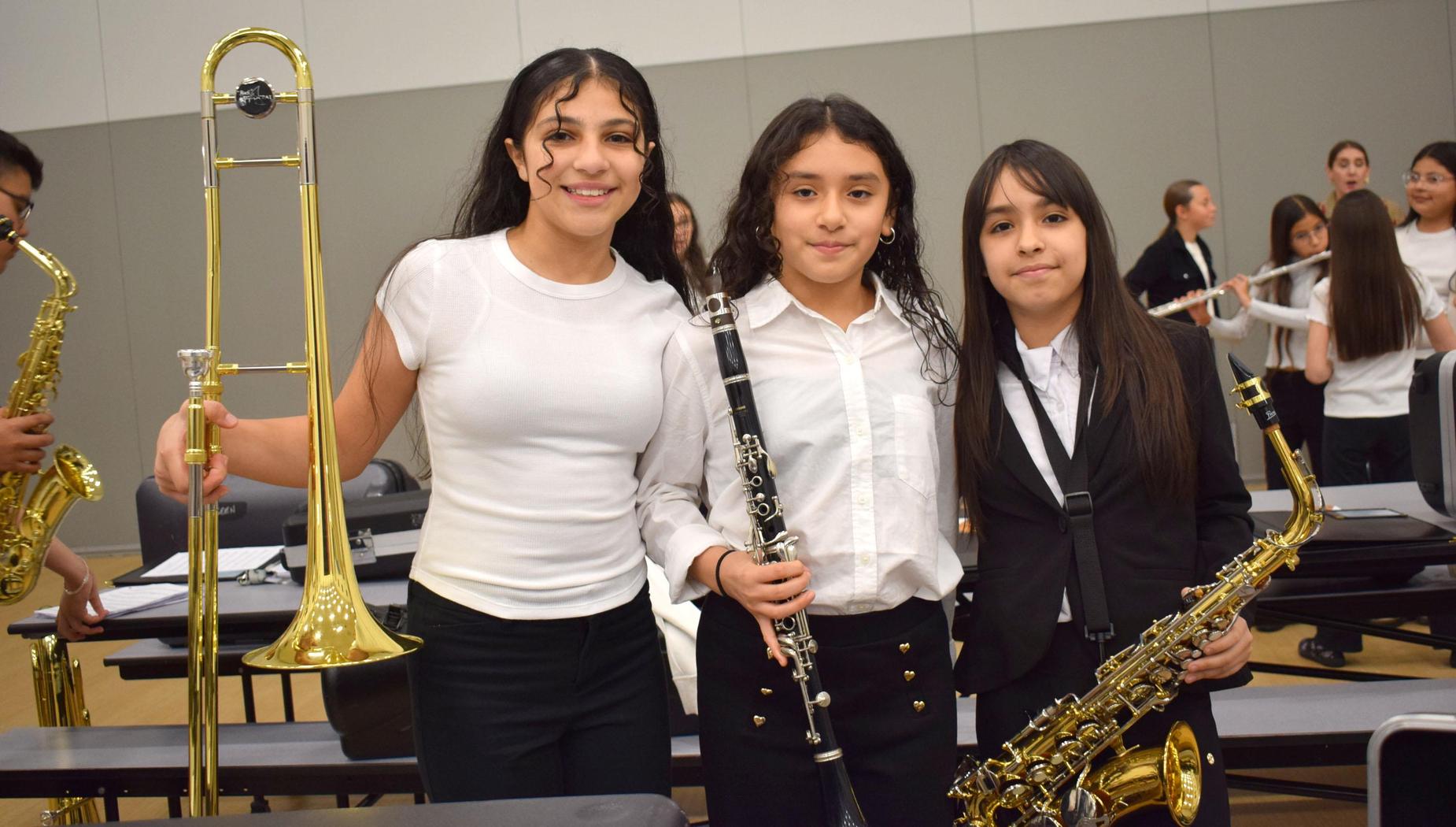 Three young musicians, one with a trombone, stand smiling in a music room.