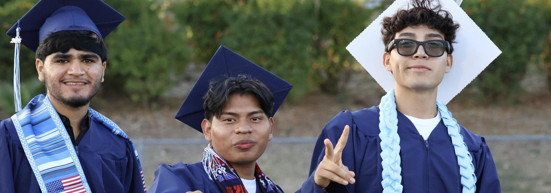 Three graduates in caps and gowns celebrating outdoors.