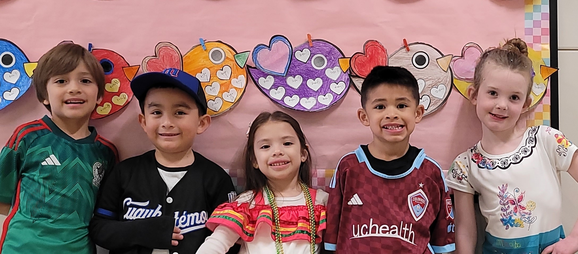 Five children smiling together in front of a colorful heart-themed backdrop.