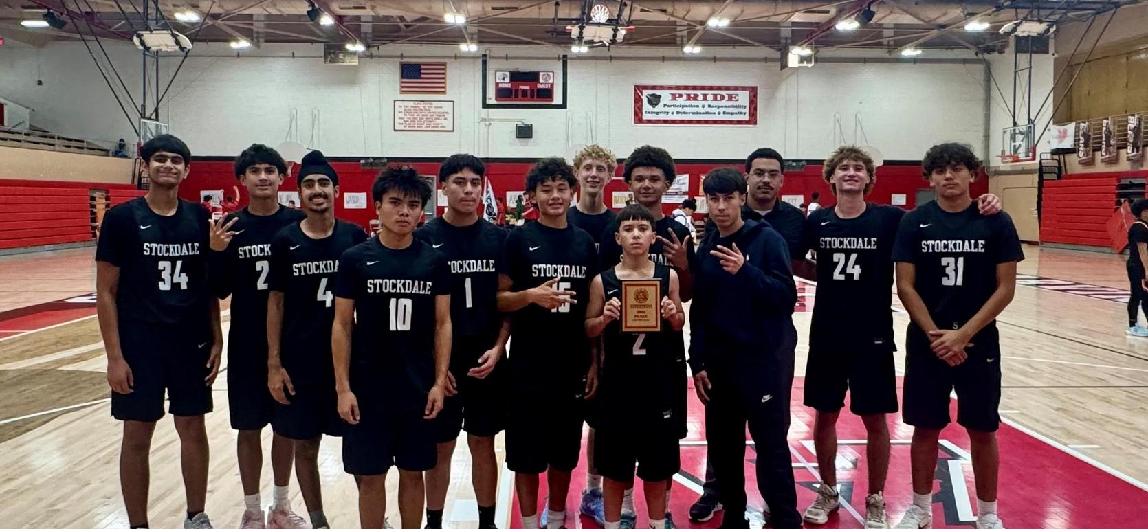 A basketball team in black jerseys, holding a trophy in a gymnasium.