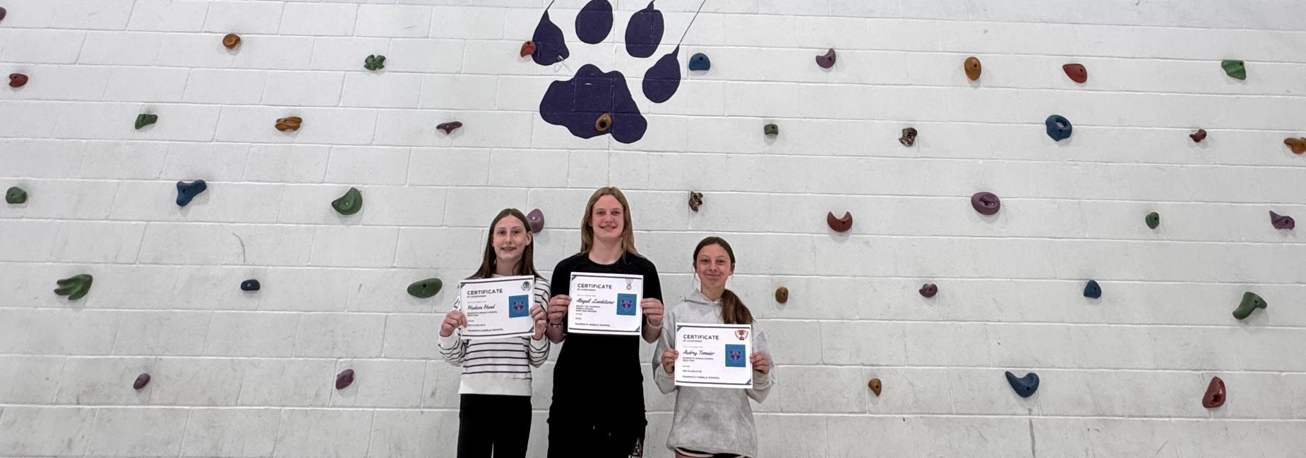 Three girls standing in front of a 'Mountain Lions' mural with rock climbing features.