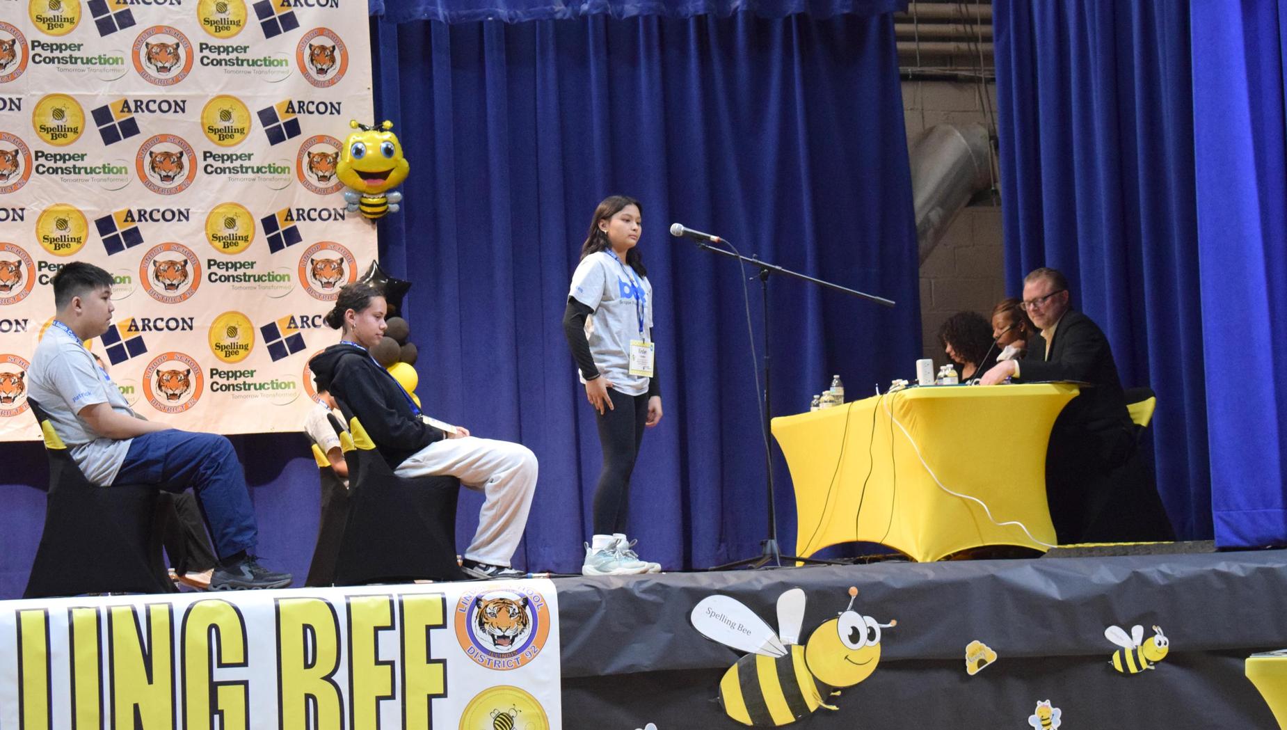 A girl stands at a microphone during a spelling bee, with judges nearby.