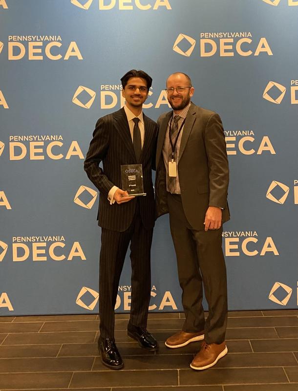 Image of a student and his advisor standing in front of a backdrop that says DECA