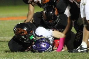 Two football players tackle an opponent wearing a purple helmet on the grass.