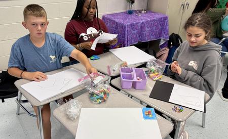 Three students sitting at desks working on art projects with various supplies.