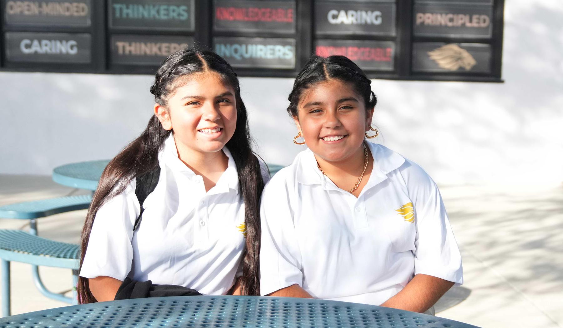two smiling girls in SJLA uniform shirts sitting at a blue picnic table.