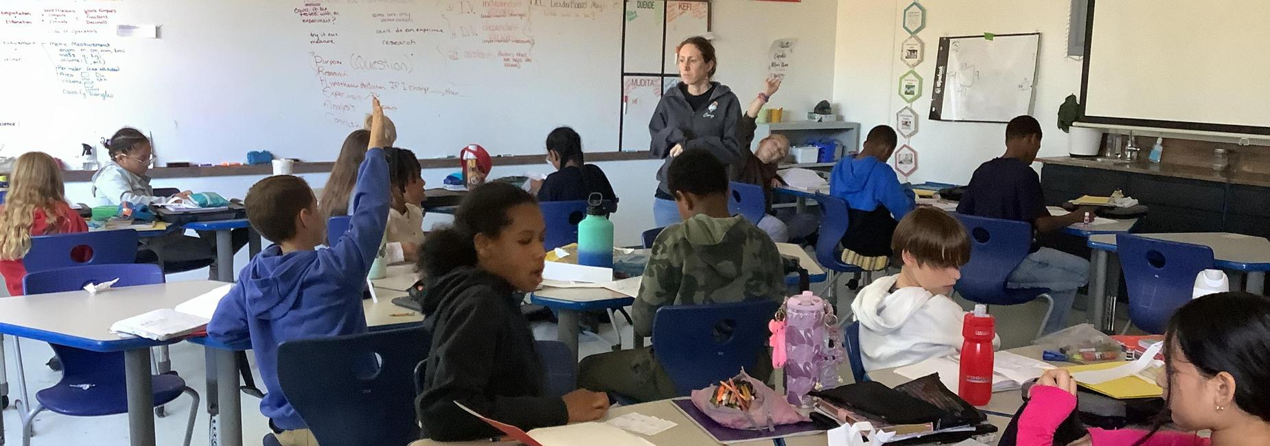 Classroom scene with students engaging, teacher standing, and various school supplies visible.