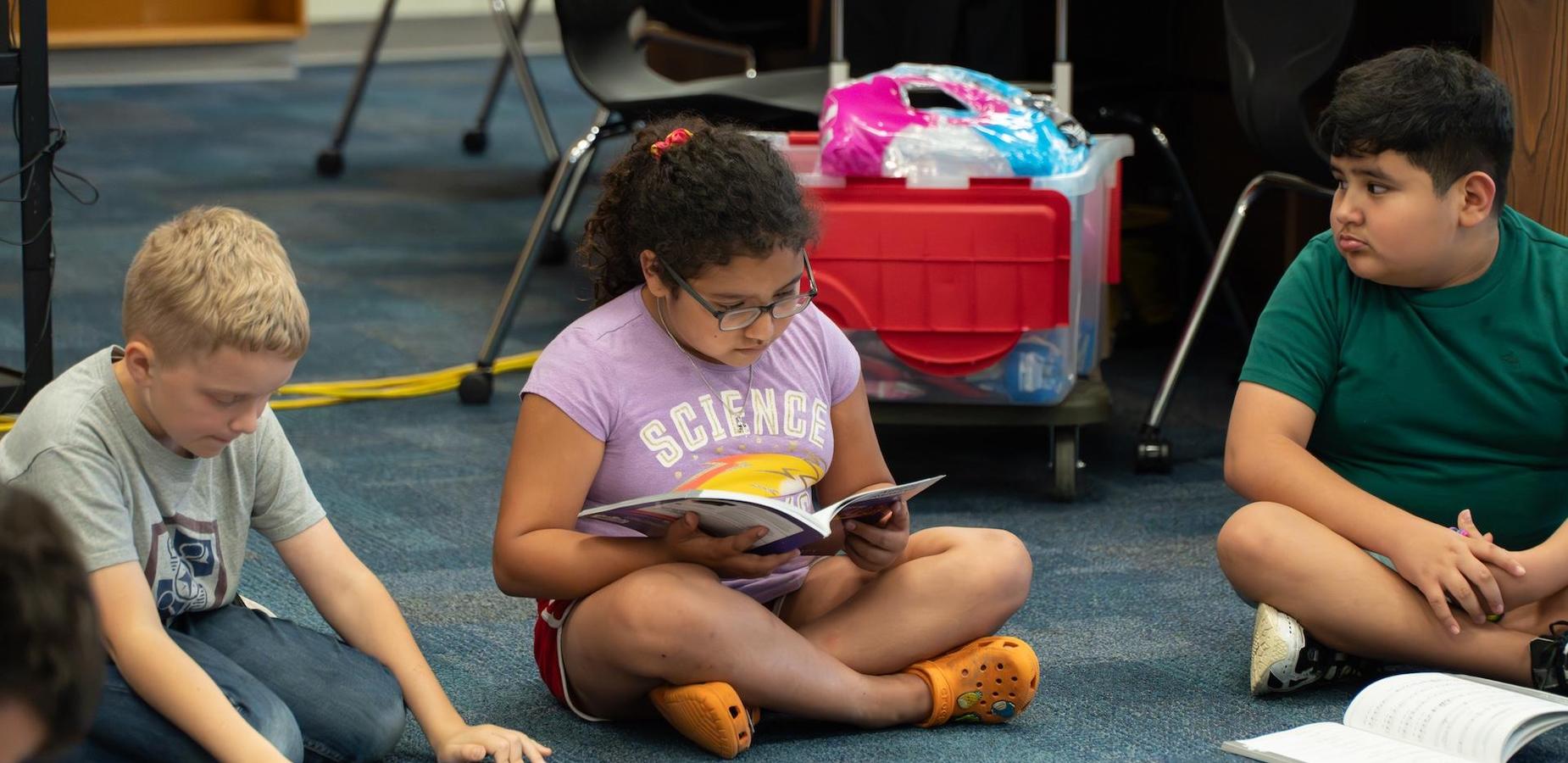 Three children sitting on the floor reading books in a classroom setting.
