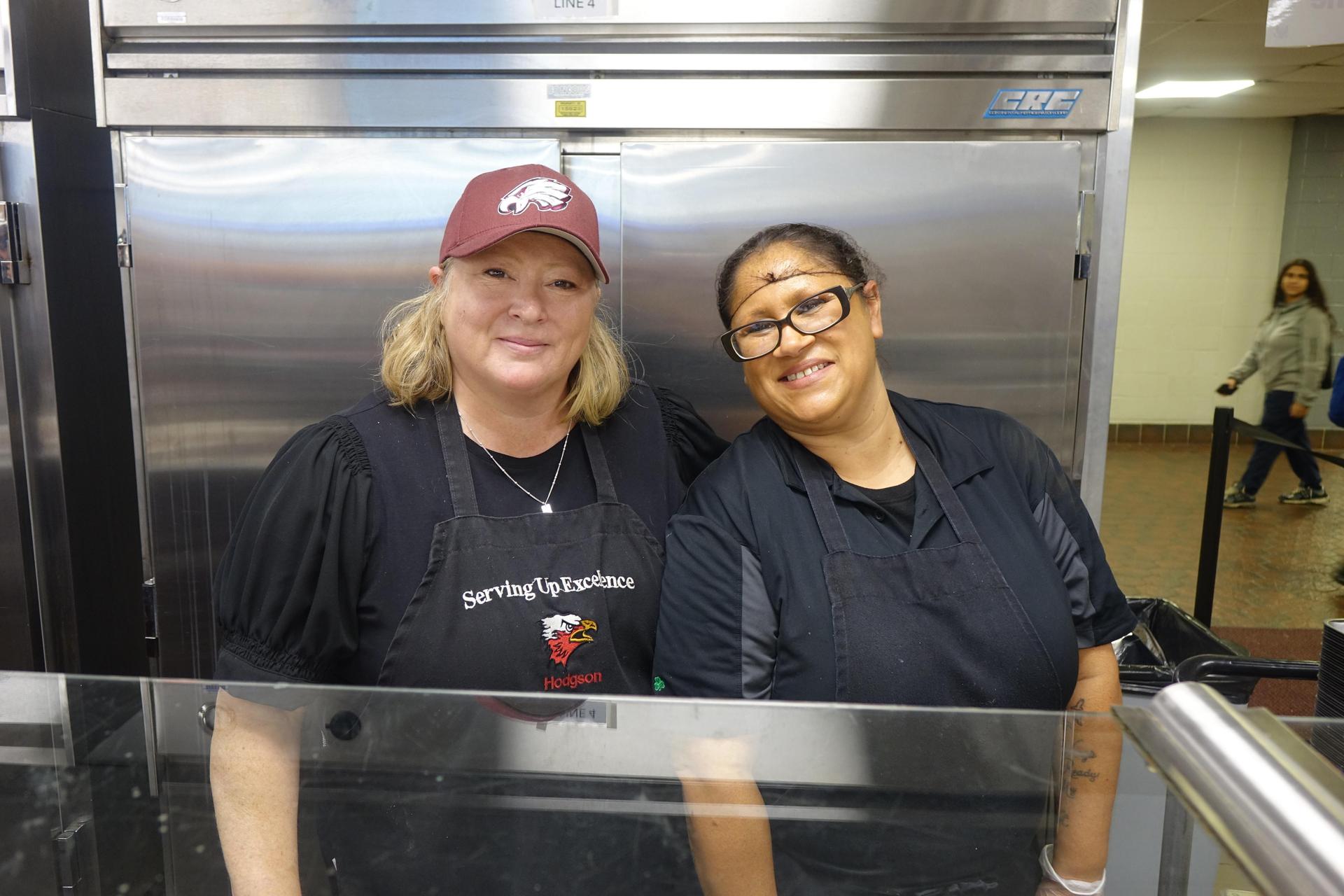 Two women in black aprons smiling behind a food service counter.