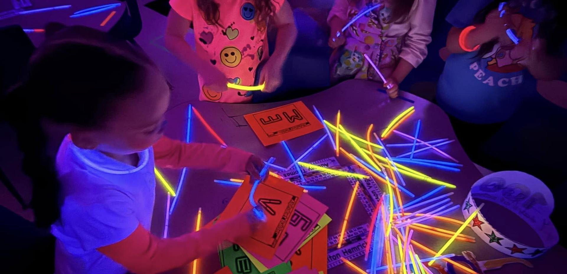 Children writing on neon cards with glow sticks in a dark room.