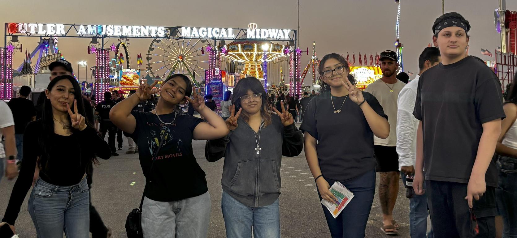 Five friends posing with peace signs at a lively fairground with lights and attractions.