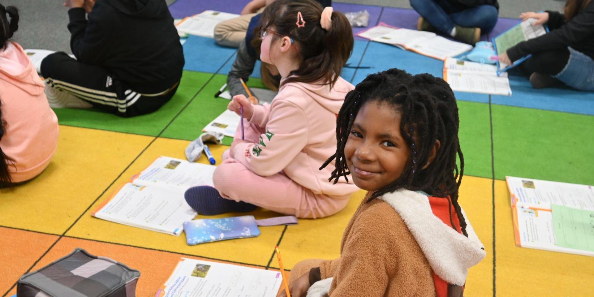 A young girl smiles while sitting on a colorful classroom floor with classmates.