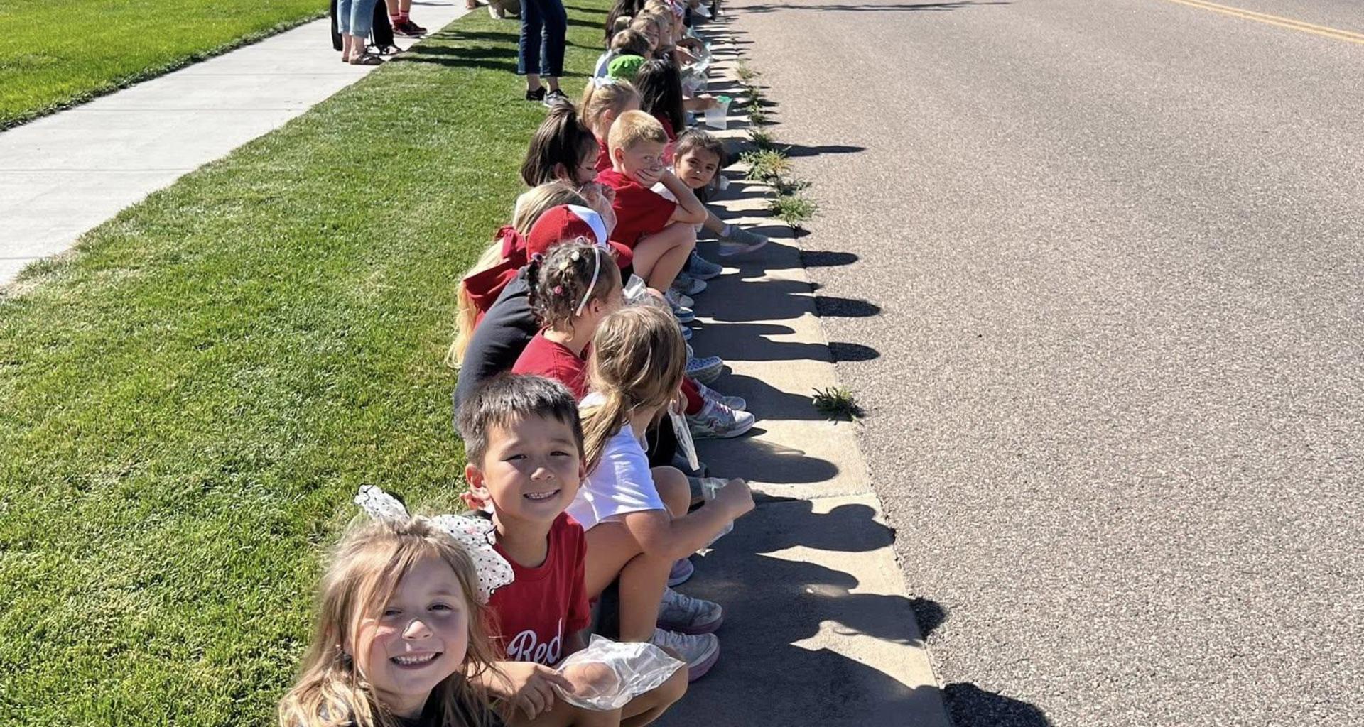 Children sitting in a line on the sidewalk by a road, smiling and chatting.