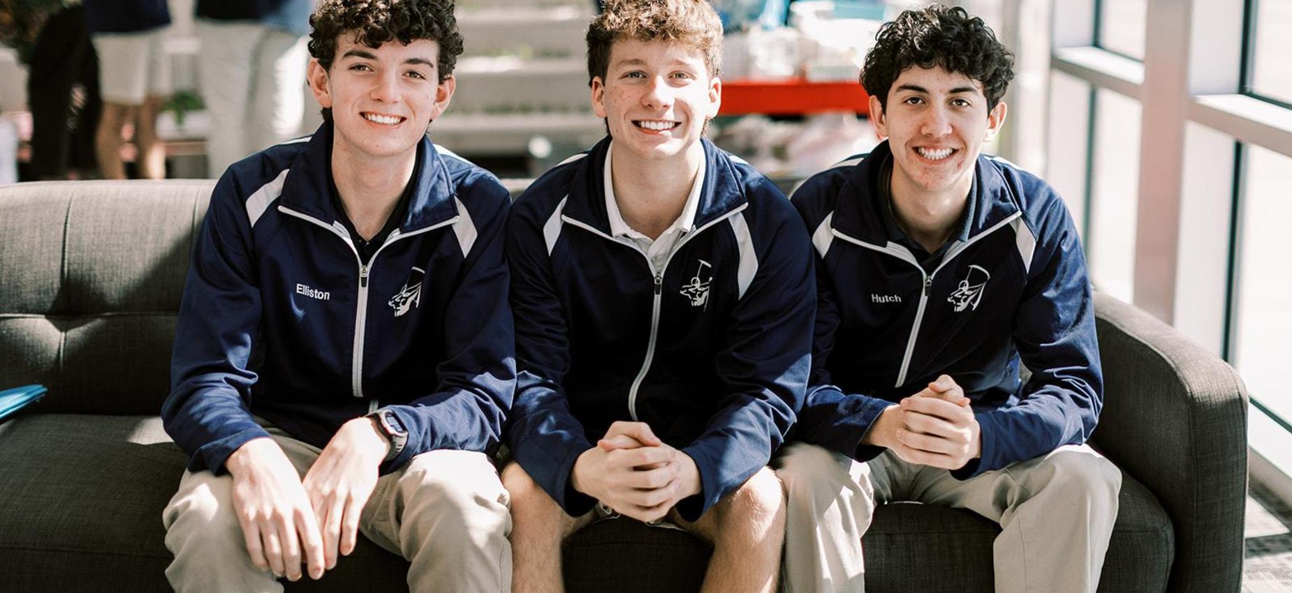 Three smiling high school students in Archers jackets sit together on a couch