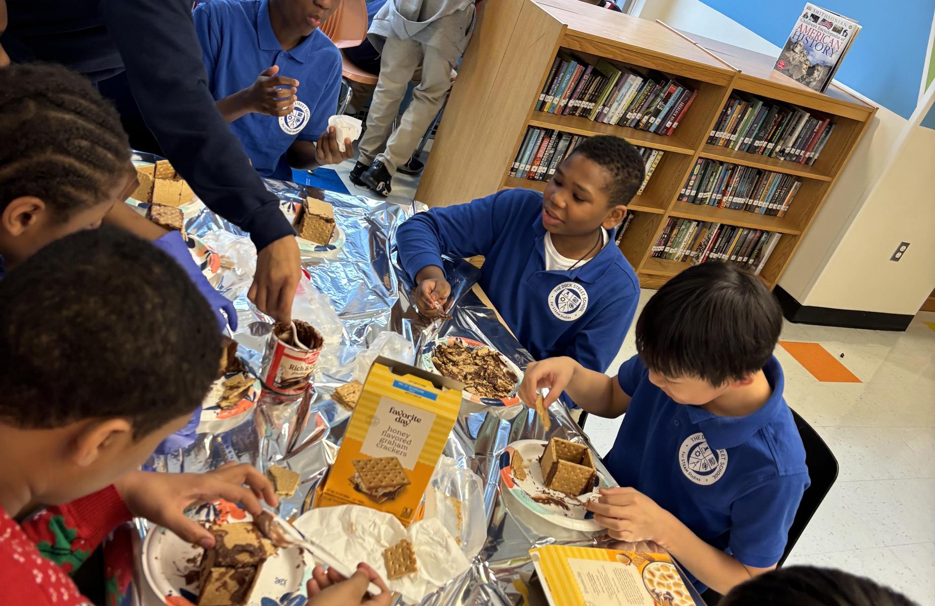 Children in blue shirts making snacks at a table in a library.