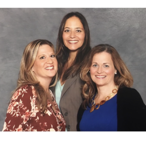 Three women posing together in professional attire against a gray backdrop.