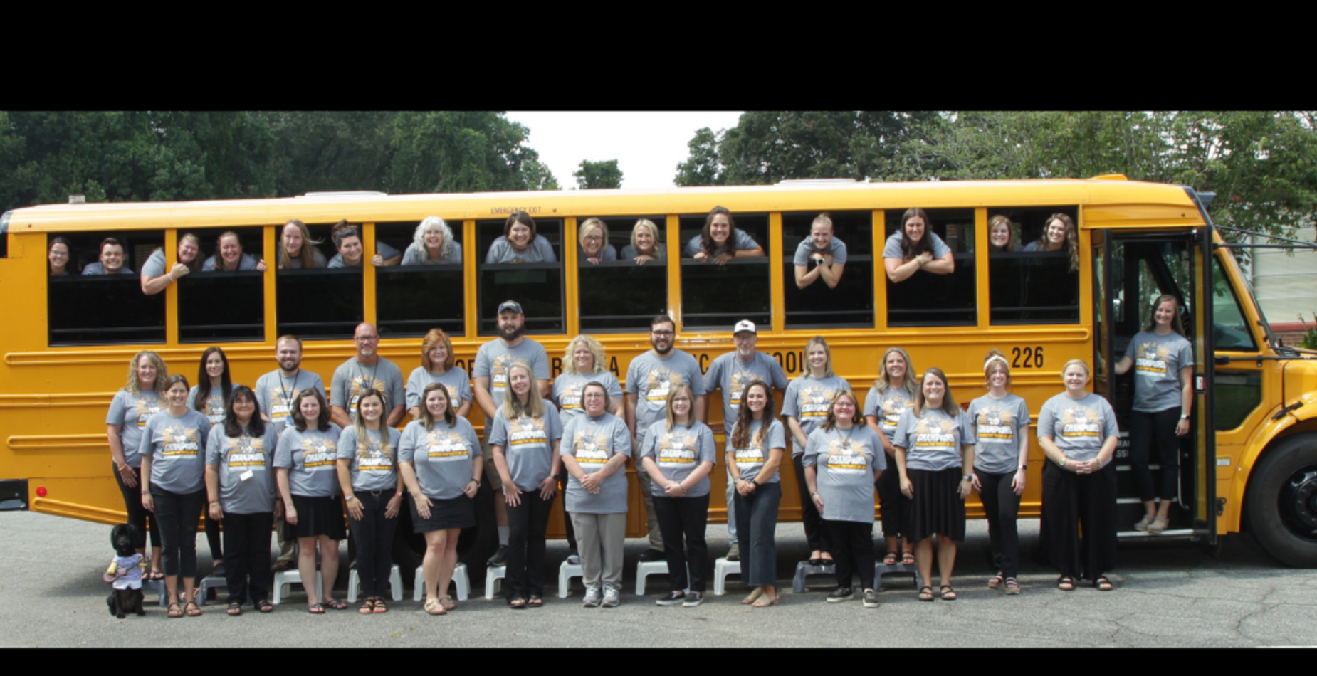 A large group of people wearing matching gray shirts poses in front of a yellow school bus.