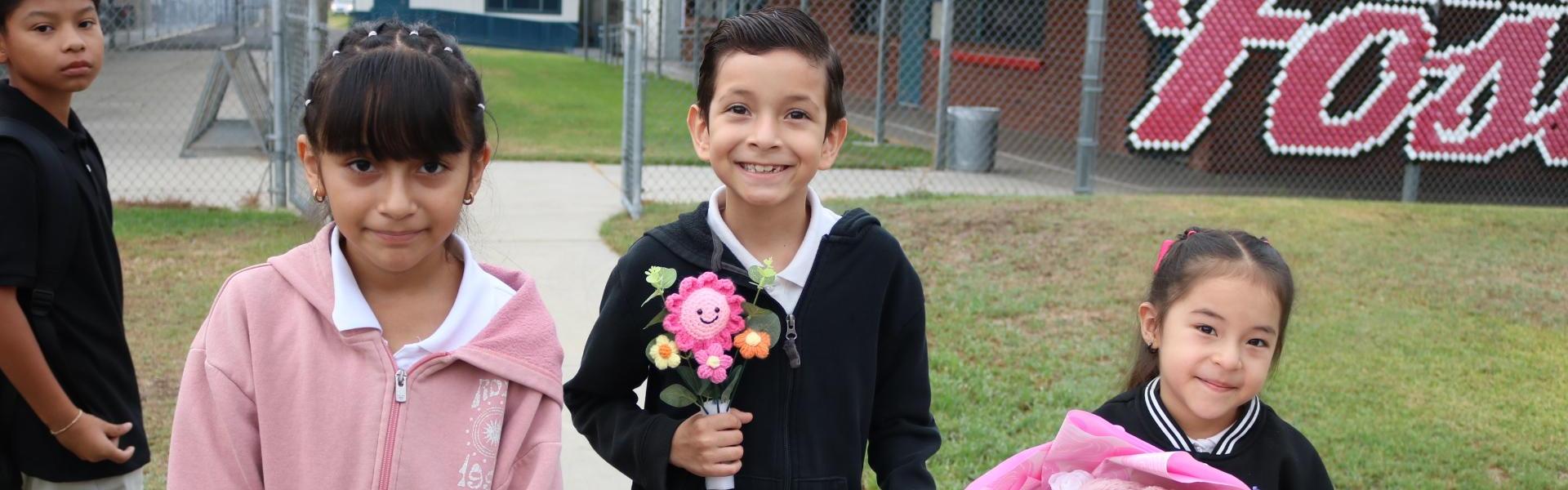 Three children smiling, one holding flowers and a teddy bear.