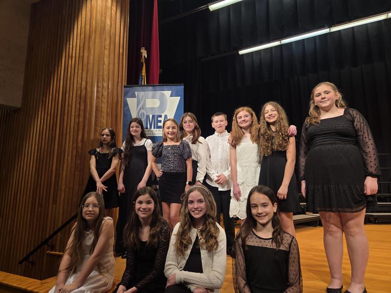 students dressed in black and white sit on edge of stage with PMEA sign behind them